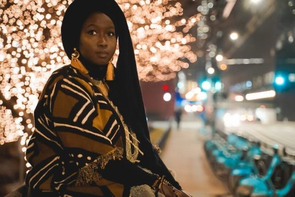 Night portrait of a female-presenting person with dark skin and long dark hair backlit by a tree with thousands of lights on it