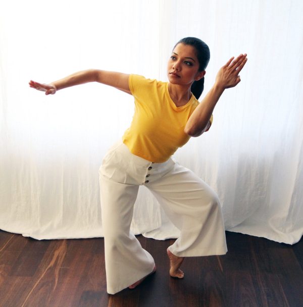 A woman dressed in a yellow top and wide white pants leans to her left with her leg bent. Her right arm is extended in a snake-arm like position. Her left arm is bent up towards her face.