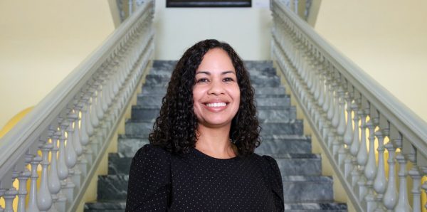 A three-quarter portrait of a smiling female-presenting person with medium-length curly black hair at the base of a marble staircase