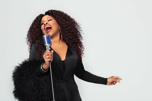 Chaka Khan dressed all in black singing into a microphone against a white background