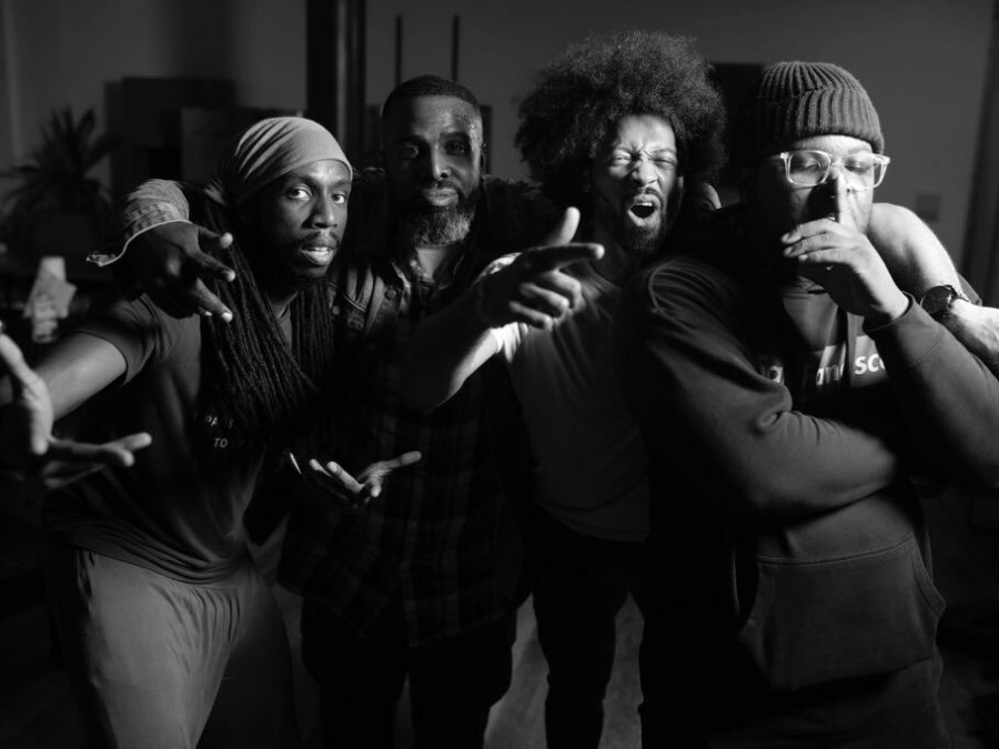 Black-and-white photo of four men posed with arms around each others' shoulders. The man closest to the camera holds his left hand in front of his mouth as if beatboxing.