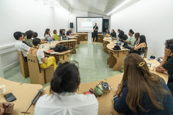 A group of young people sit at desks facing a projector screen at the front of a room.