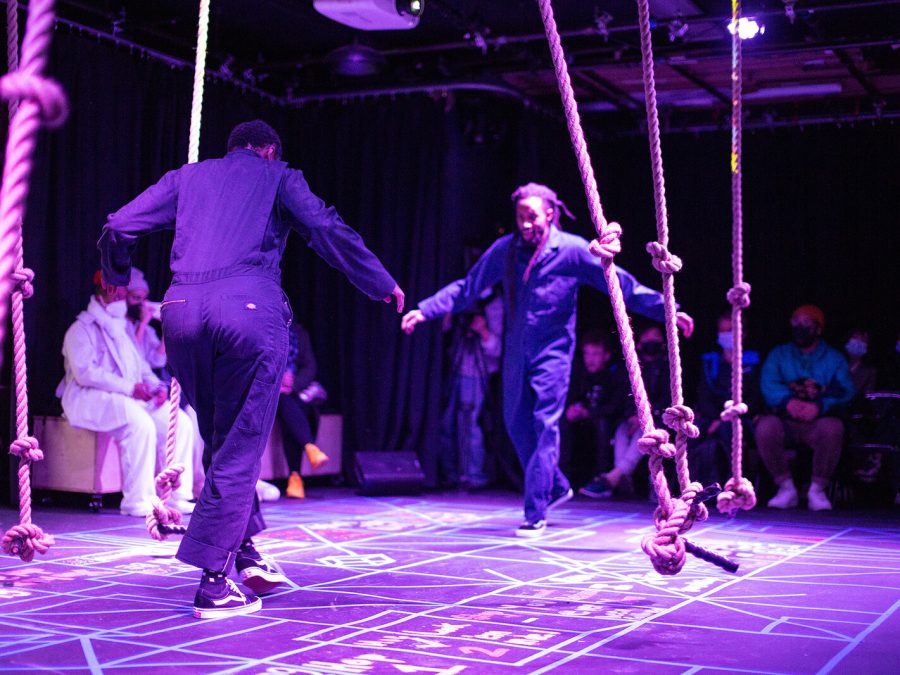 Two people facing each other among a purple-lit space full of thick, knotted ropes hanging down.