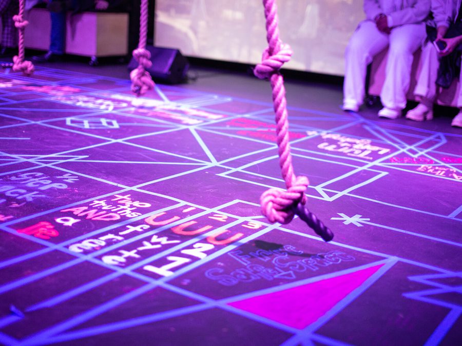 A thick rope with notes hangs above a purple-lit floor covered in diagrams.