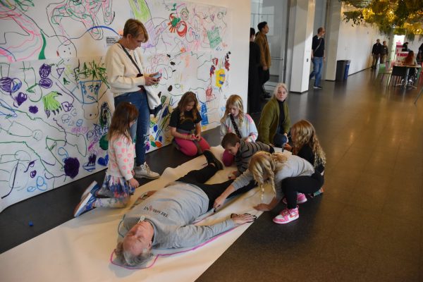 A group of people gather around an adult lying on a large piece of paper. Some of them trace the outline of their body,