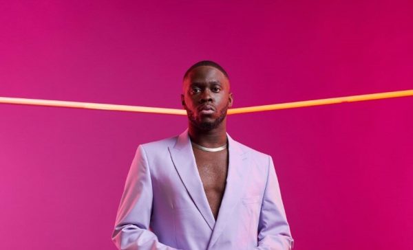 A man in a lavender suit stands in a magenta-colored room.