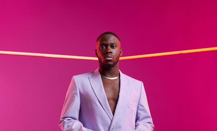 A man in a lavender suit stands in a magenta-colored room.