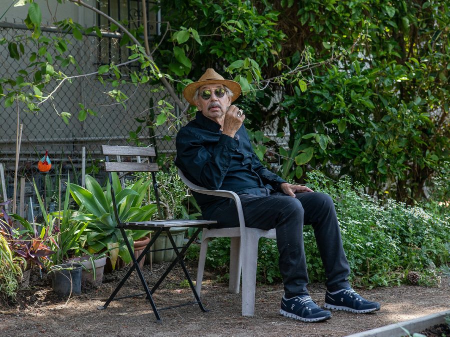 Artist David Lamelas sits on a plastic chair surrounded by green foliage.