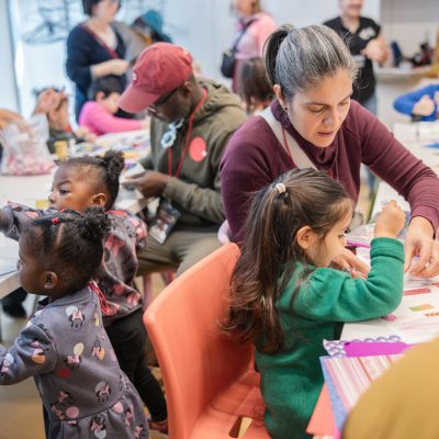Kids and adults sit at various tables engaged in artmaking activities