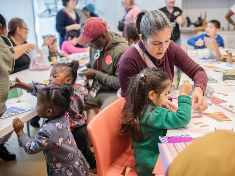 Kids and adults sit at various tables engaged in artmaking activities