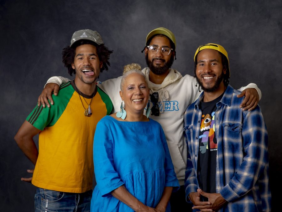 A group portrait of a woman surrounded by three younger men against a gray backdrop.