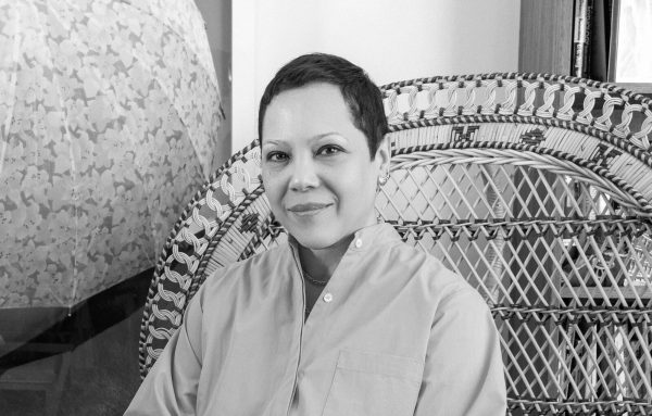 Black and white photo of a female presenting person with short hair poses in a peacock chair with an artwork hanging to their right