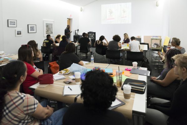 A large group of people sit at various tables filled with notebooks, pens, paper, and coffee cups in a small white room