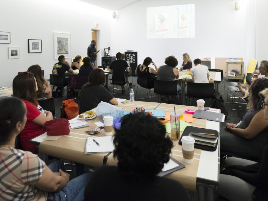 A large group of people sit at various tables filled with notebooks, pens, paper, and coffee cups in a small white room
