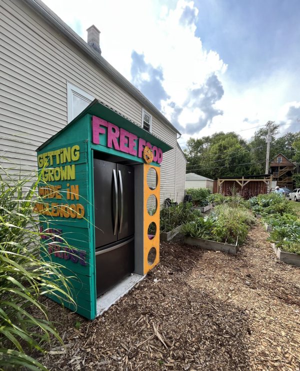 A colorful structure with a black fridge inside in a garden setting. Pink letting on top declares Free Food