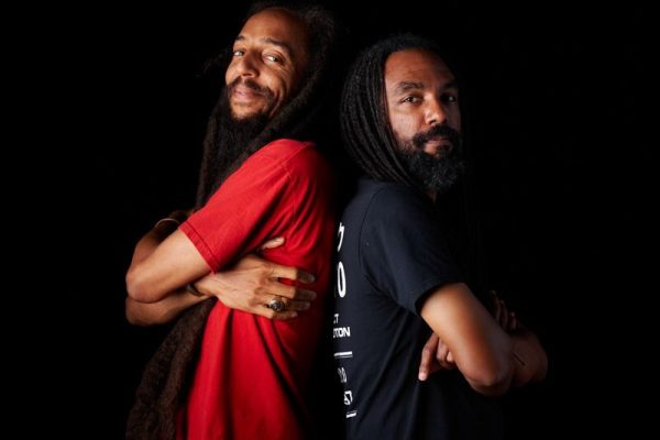 Two male-presenting people with medium brown skin pose back to back against a black background