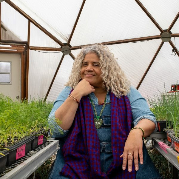 A female-presenting person with white curly hair sits in a greenhouse, their right hand resting under their chin