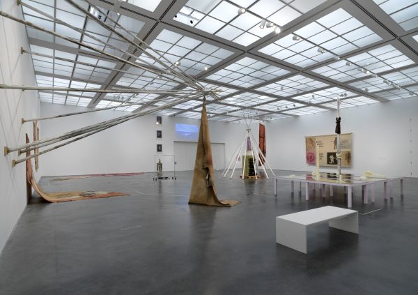 A large gallery space with many artworks installed on the walls and floor, including posts put together in a tipi formation that juts out from the wall.