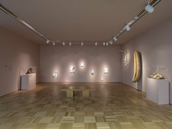A pink painted gallery space with a wooden checkerboard floor. Spotlighted artworks hang on the walls.