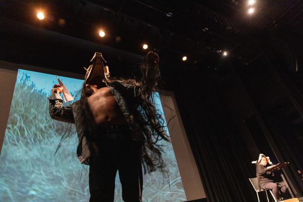 View up at a person dressed in a furry coat that is open to expose a bare chest. They wear a cardboard mask and hold a paper airplane in their right hand.