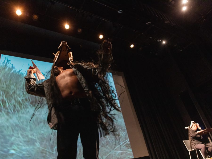 View up at a person dressed in a furry coat that is open to expose a bare chest. They wear a cardboard mask and hold a paper airplane in their right hand.