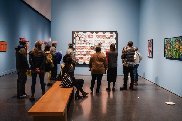 A group of people gather around a large painting in a blue gallery filled with other artworks.