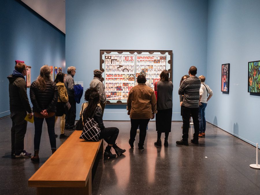 A group of people gather around a large painting in a blue gallery filled with other artworks.