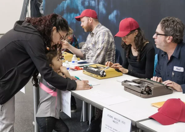 people sit waiting at a desk with typewriters as an adult and a child lean over to fill out paperwork