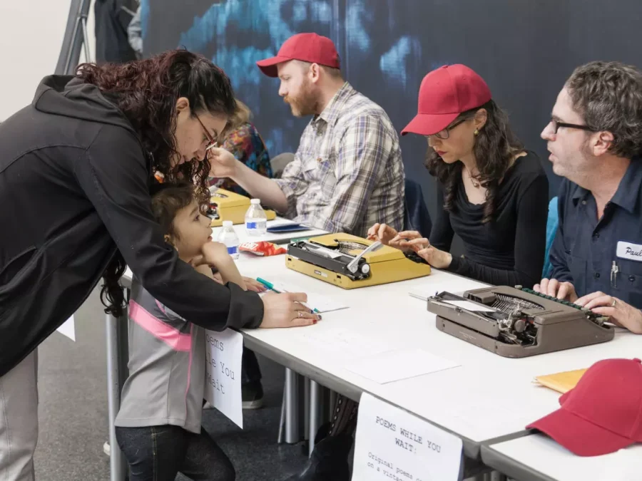 people sit waiting at a desk with typewriters as an adult and a child lean over to fill out paperwork