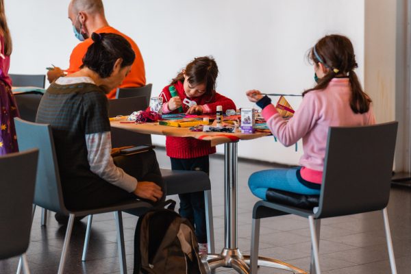 Two children and an adult sit around a square table deeply focused on an art project.