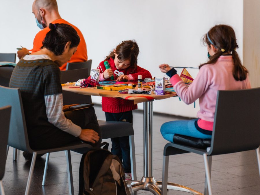 Two children and an adult sit around a square table deeply focused on an art project.