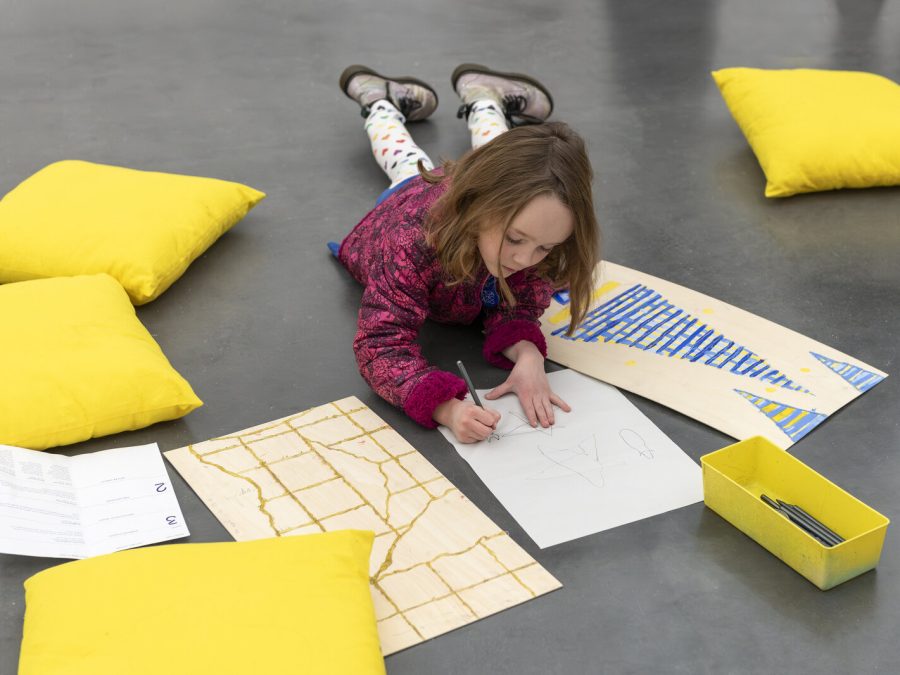 A child lays on a concrete floor and colors while surrounded by yellow pillows and sheets of paper
