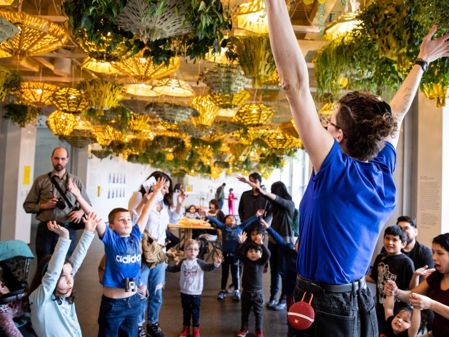 A group of kids raise their arms, mirroring the pose of an adult standing in front of them in a room full of planters hanging from the ceiling