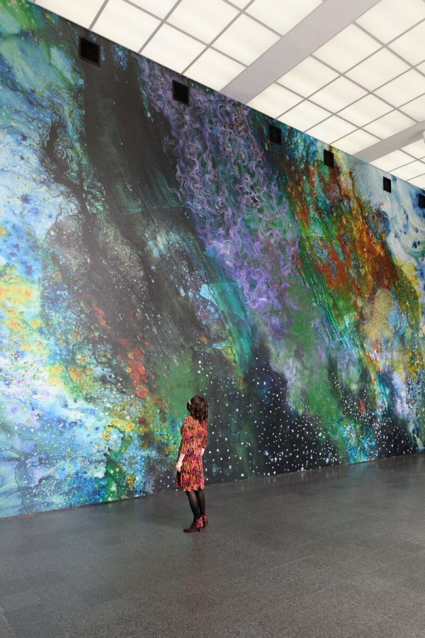 A person stands in a two-story space gazing at a colorful abstract mural on the two-story wall