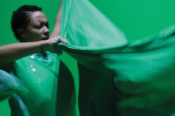 A Black person with curly hair wearing a green costume grasps the edge of a piece of green fabric.