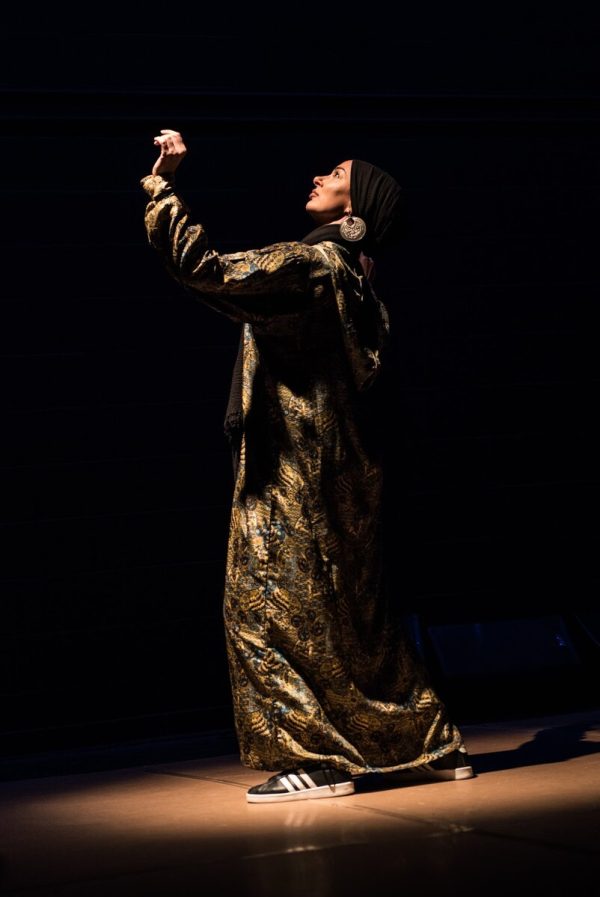 Full-length portrait of a person in side profile wearing a scarf that covers their hair and a long robe against a black background. They look up towards a spotlight, raising their left hand towards it.