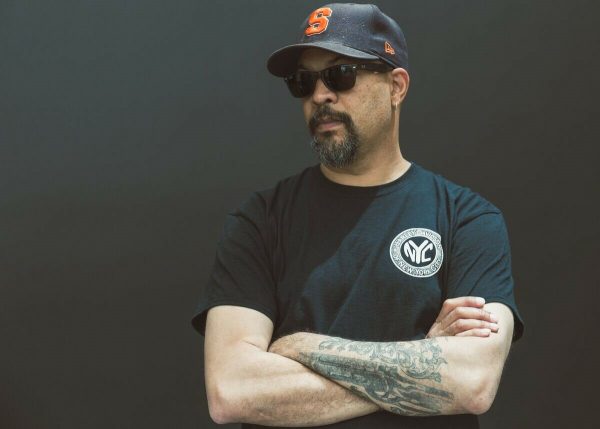 A man wearing a black, short-sleeved t-shirt and black hat poses against a plain, dark wall. His tattooed arms are crossed.