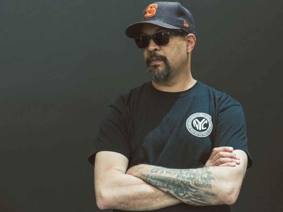 A man wearing a black, short-sleeved t-shirt and black hat poses against a plain, dark wall. His tattooed arms are crossed.