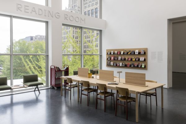 A room labeled Reading Room over large, square windows. It features a long table with lights and dividers and a wall full of books displayed with their covers facing outwards.