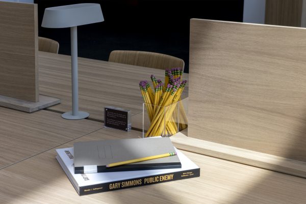 Closeup of a light wood desk with wooden privacy screens, and lamp, and a container of pencils in the middle. Close to the edge of the frame are two books and a pencil resting on top.