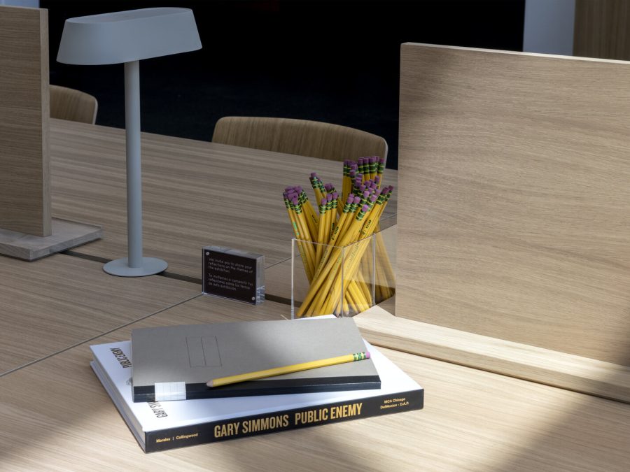 Closeup of a light wood desk with wooden privacy screens, and lamp, and a container of pencils in the middle. Close to the edge of the frame are two books and a pencil resting on top.