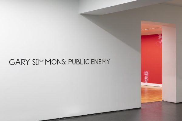 A sliver of a room painted red with white drawings on it is just visible beyond a wall with black text that reads Gary Simmons: Public Enemy.