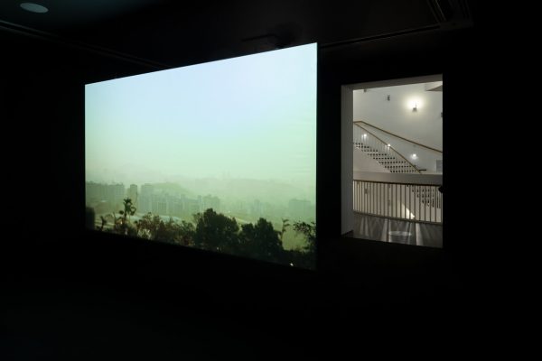 A film plays on a wall in a darkened room next to a doorway revealing a brightly lit staircase.