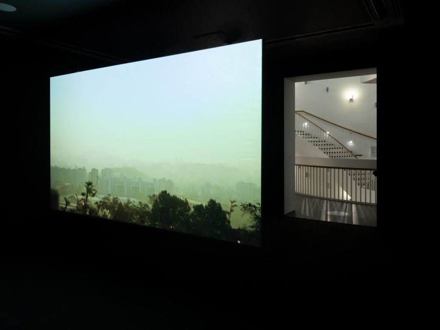 A film plays on a wall in a darkened room next to a doorway revealing a brightly lit staircase.