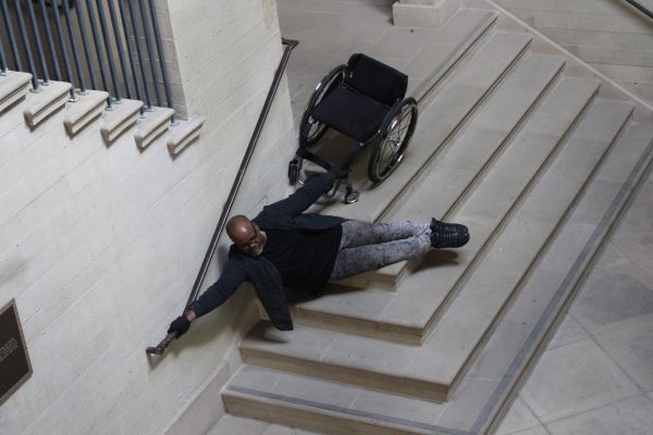 A Brown-skinned large-bodied black person with shaved head & beard, wearing glasses, black shirt and grey jeans, with one hand on a rail and the other holding a wheelchair, seemingly floating above a set of stairs.