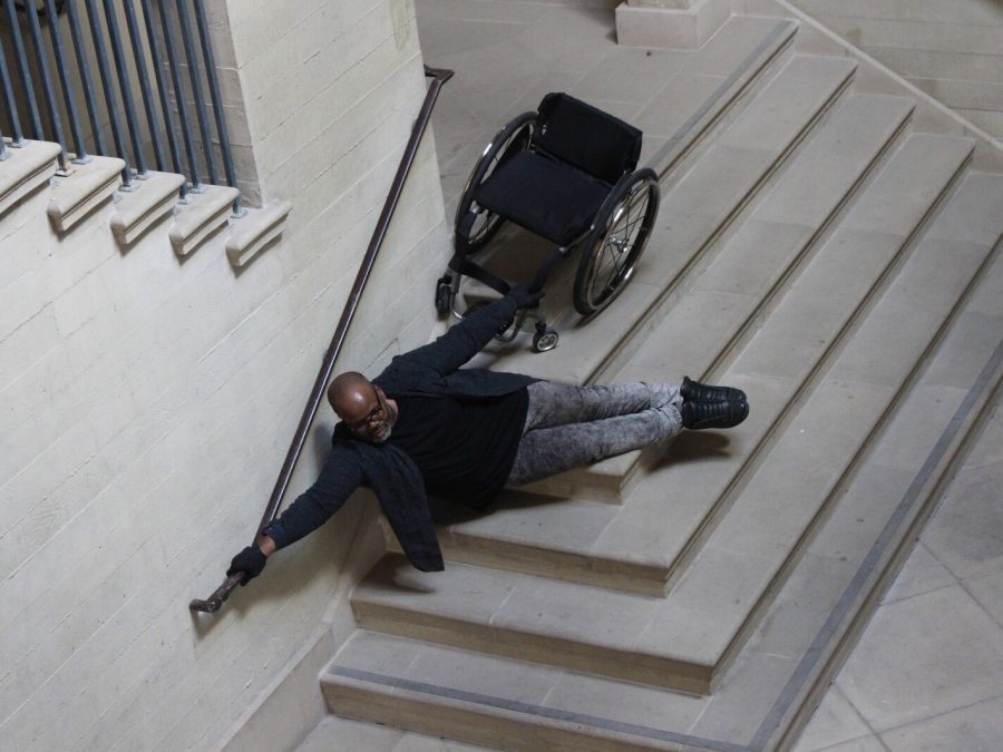 A Brown-skinned large-bodied black person with shaved head & beard, wearing glasses, black shirt and grey jeans, with one hand on a rail and the other holding a wheelchair, seemingly floating above a set of stairs.