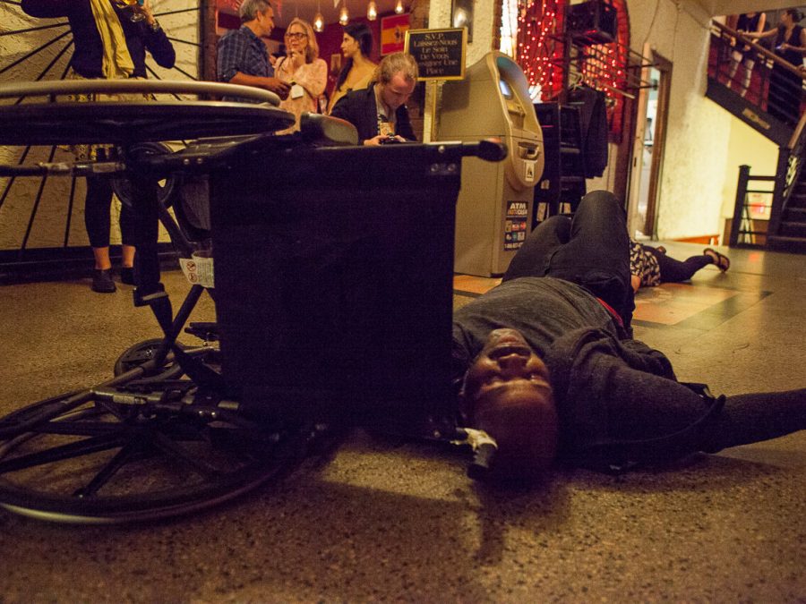On the landing of a staircase, a brown-skinned large-bodied black person lies on a marble floor next to a manual wheelchair that is turned on its side. In the background is a automated bank machine and a diverse group of people in various positions. Some are looking intently at the person, others are talking with each other.