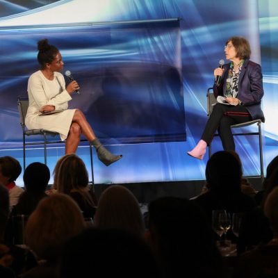 Two women sit on a stage in front of a crowd of people.