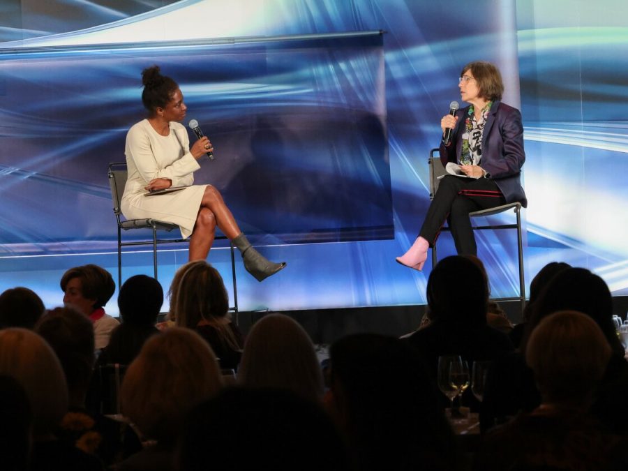Two women sit on a stage in front of a crowd of people.