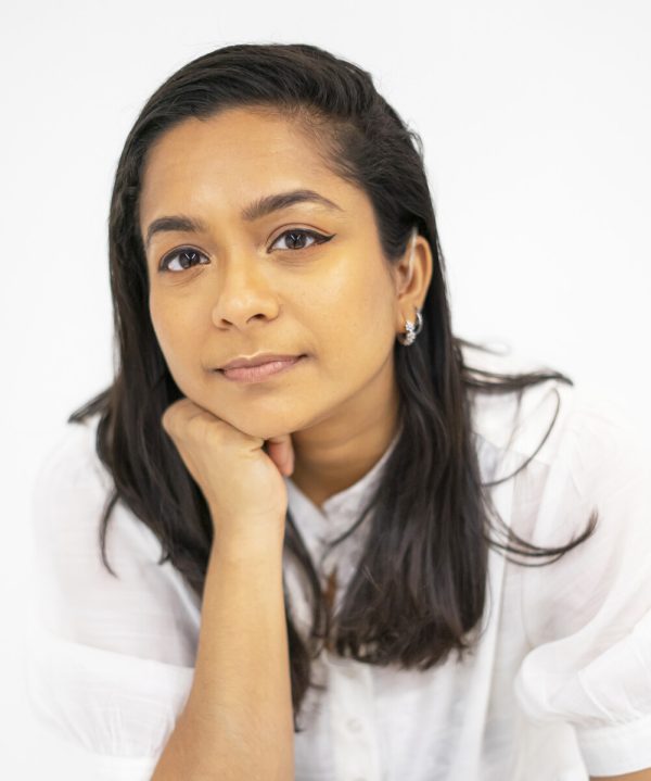 Portrait of a person with medium-length, dark brown hair dressed in white against a white background. Their chin rests atop a clasped right hand.